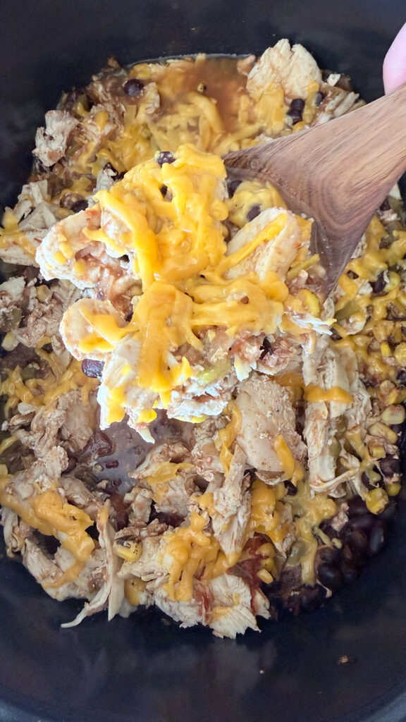 cooked crockpot chicken burrito bowl filling on a spoon with the rest of the chicken filling in the base of the crockpot in the background