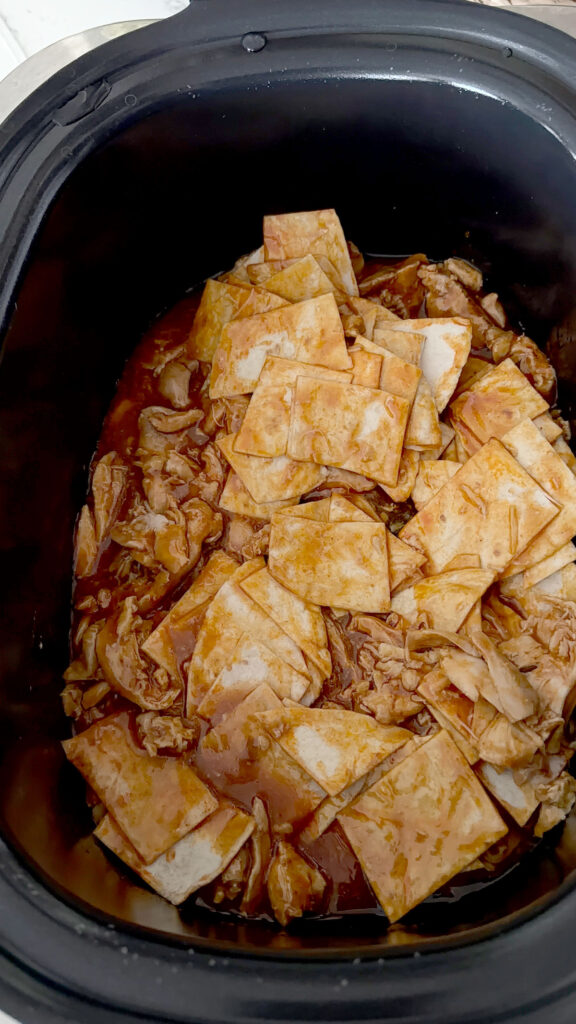 A black slow cooker bowl with shredded chicken and cut tortillas on top.