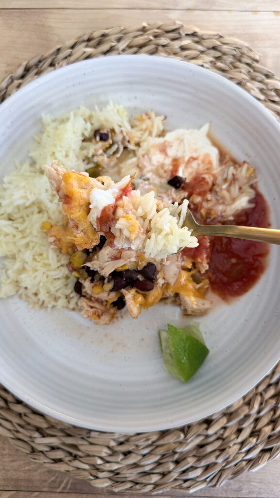 crockpot chicken burrito bowl filling on fork with plated chicken burrito bowl filling surrounded by rice, sour cream, salsa, and a lime wedge in white bowl in background on placemat