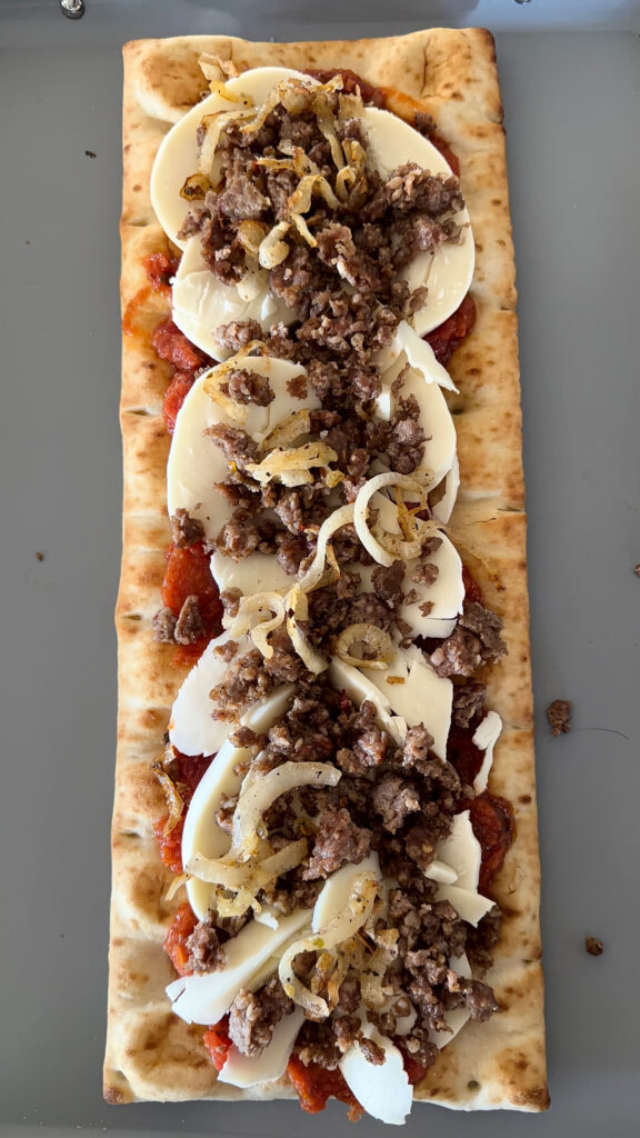 flatbread pizza crust topped with bruschetta sauce, sliced mozzarella cheese, crumbled sausage, and caramelized onions on baking sheet