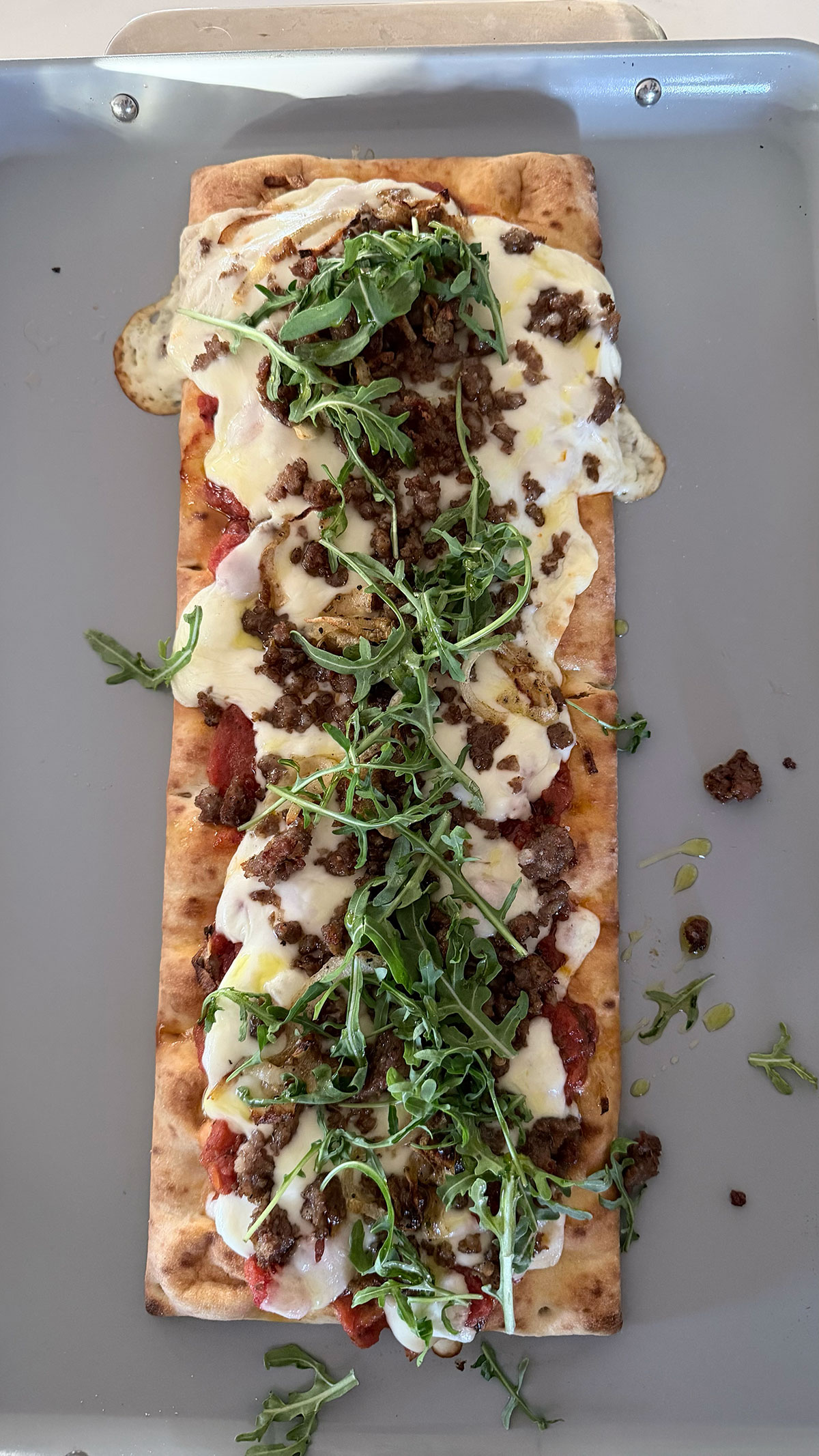 flatbread pizza crust topped with bruschetta sauce, sliced mozzarella cheese, crumbled sausage, caramelized onions and arugula, on baking sheet