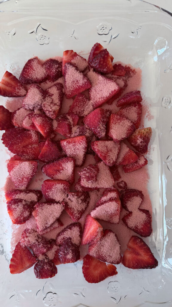Sliced strawberries at bottom of clear baking dish with strawberry jell-o powder sprinkled on top