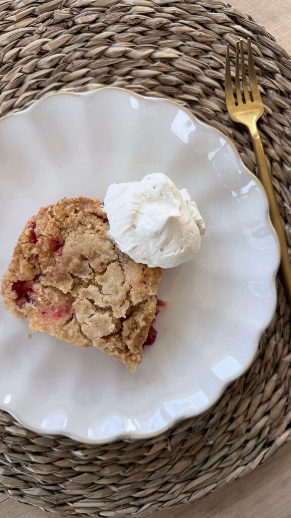 Finished and baked slice of strawberry dump cake on white plate with dollop of whipped cream. White plate on placemat with fork next to it.
