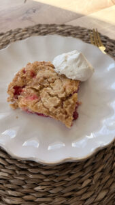 Slightly side angle of finished and baked slice of strawberry dump cake on white plate with dollop of whipped cream. Placemat and fork in background.