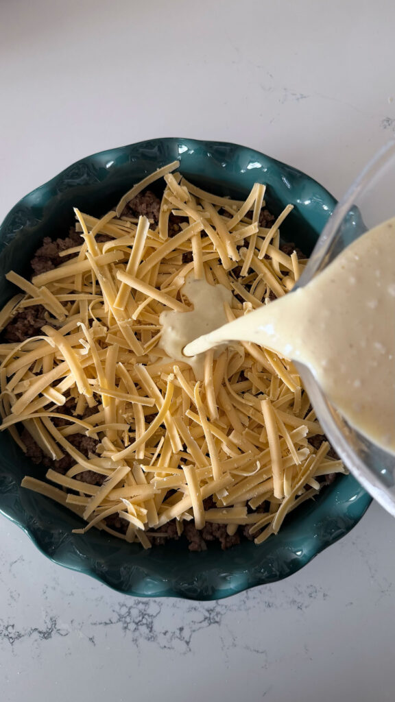 cooked ground beef topped with shredded cheddar cheese in teal pie plate with bisquick mixture being poured in, on white counter