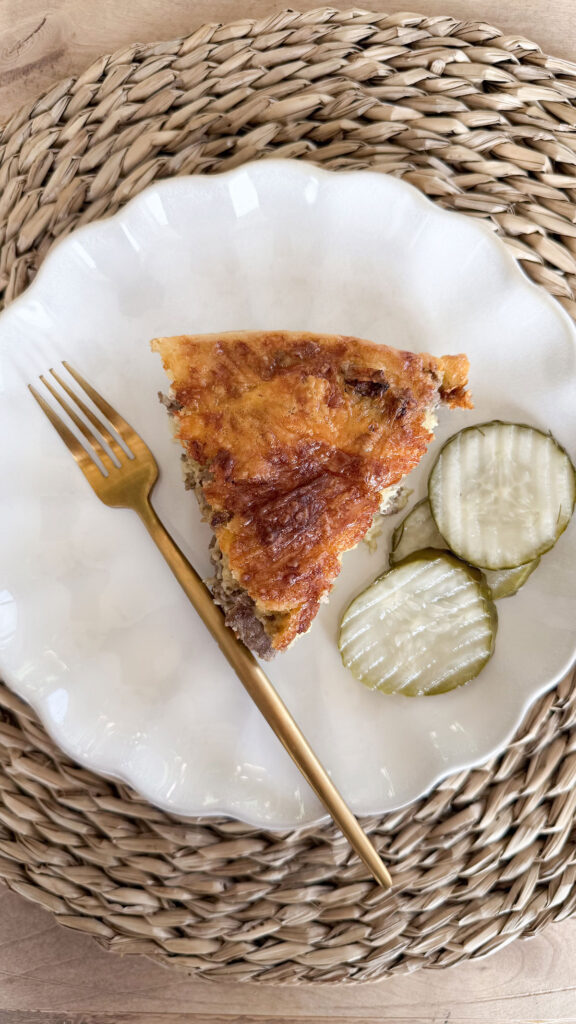 Slice of baked cheeseburger pie on white plate next to gold fork and sliced pickles. White plate on placemat.