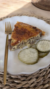 Darker version of side angle of baked cheeseburger pie slice on white plate next to gold fork and sliced pickles. White plate on placemat.
