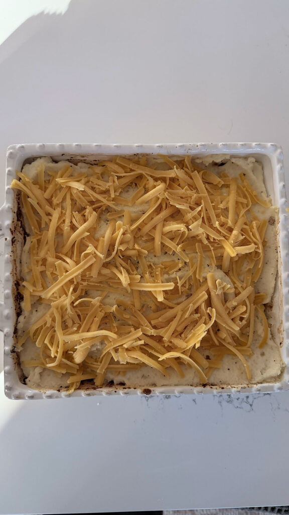 shredded yellow cheddar cheese on top of mashed potatoes and ground meat in white dish