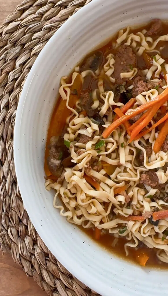 An overhead image of slow cooker beef ramen in a white bowl on a wicker placemat.