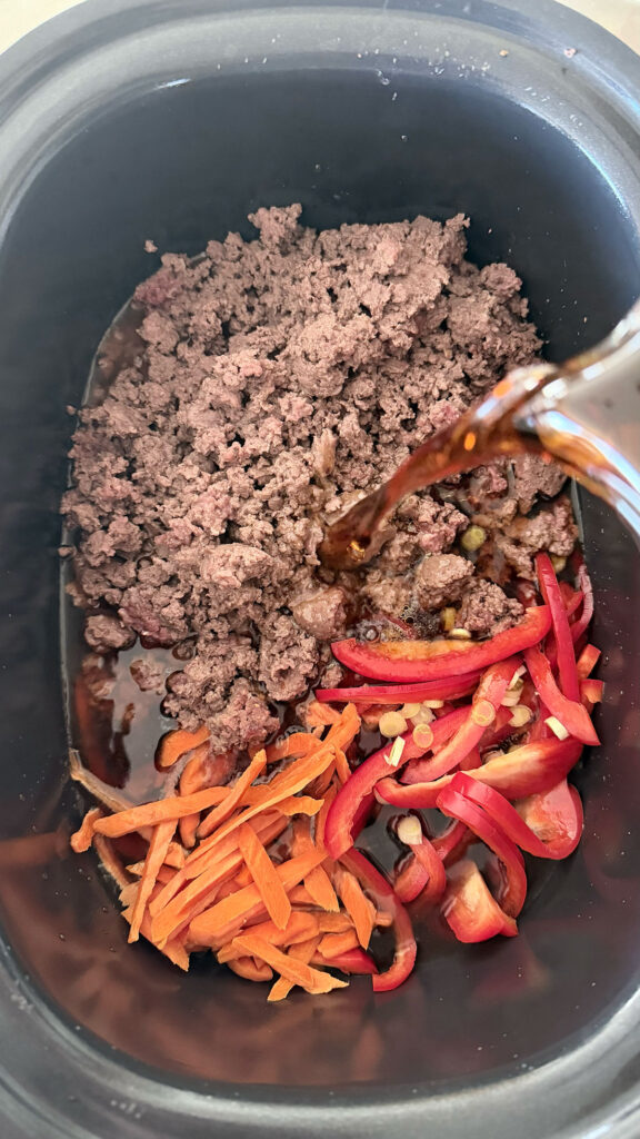 Beef broth being poured into a slow cooker bowl with ground beef, red bell pepper, and carrots.