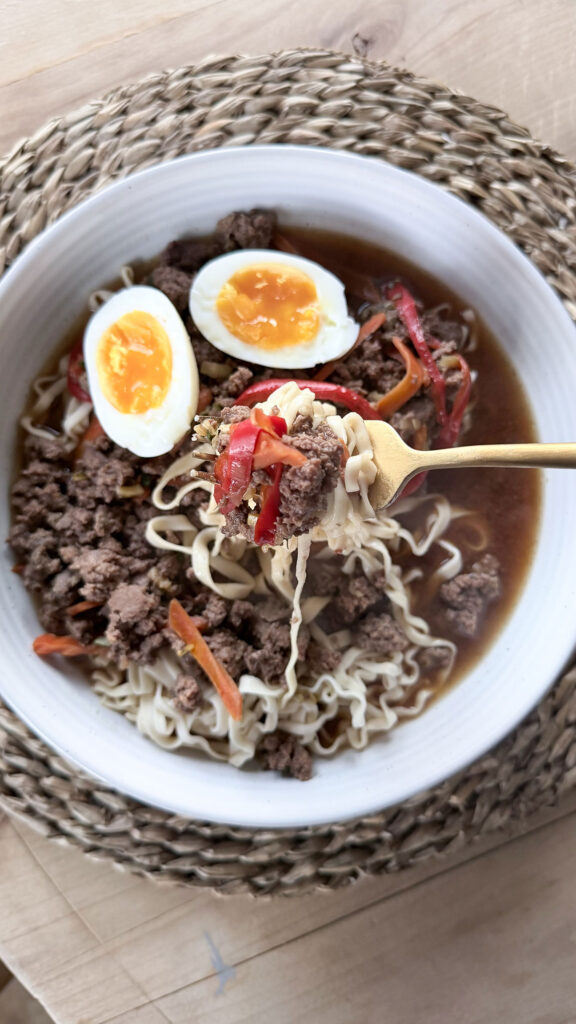 A gold fork holding up some of the slow cooker beef ramen in a white bowl on a wicker placemat.