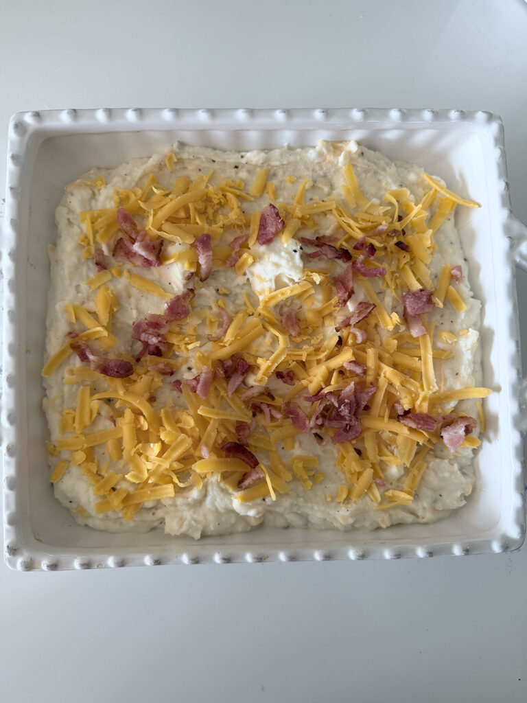 Twice baked mashed potatoes in a white baking dish with shredded cheese and bacon on top.