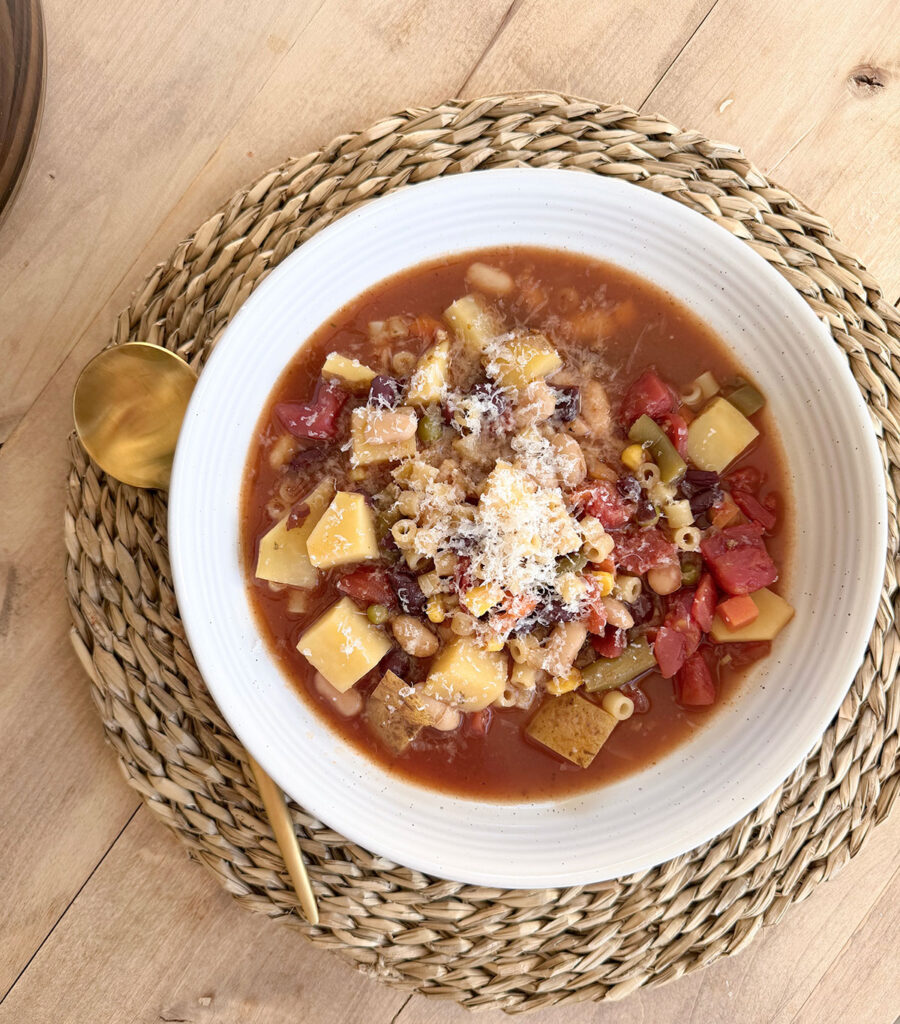 wider shot of bowl of crockpot minestrone in white bowl on placemat with gold spoon