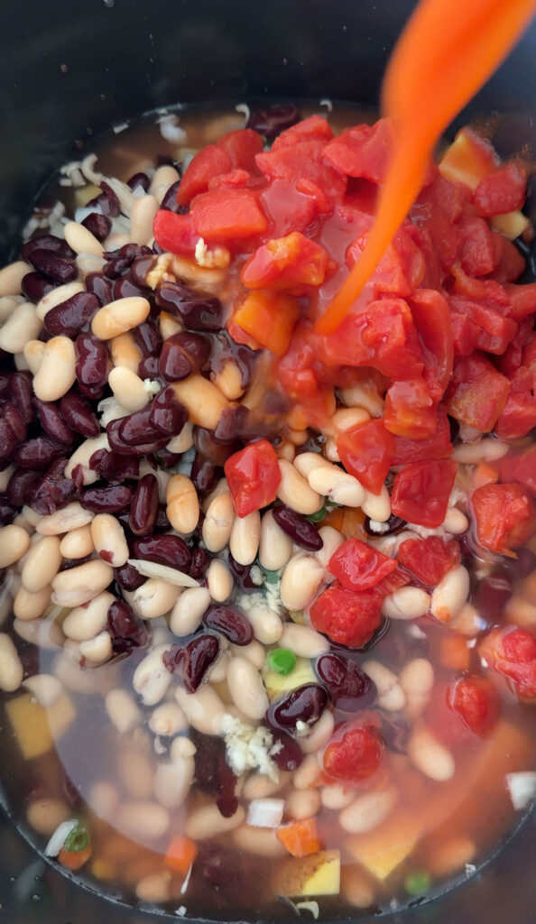 V8 being added to crockpot with canned tomatoes, beans and veggies