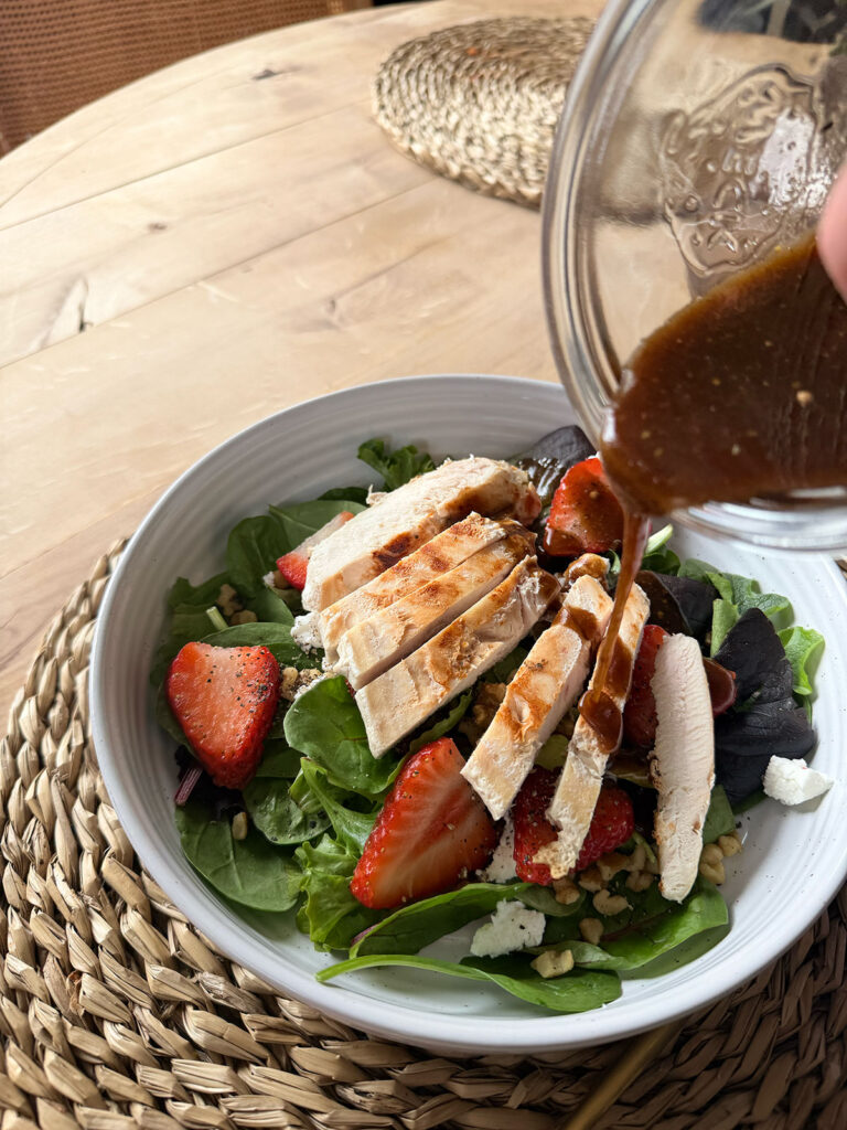 another angle of Strawberry Goat Cheese Salad in white bowl on placemat with dressing being poured over