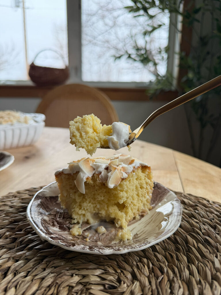 slice of coconut poke cake on plate with forkful removed, window in background