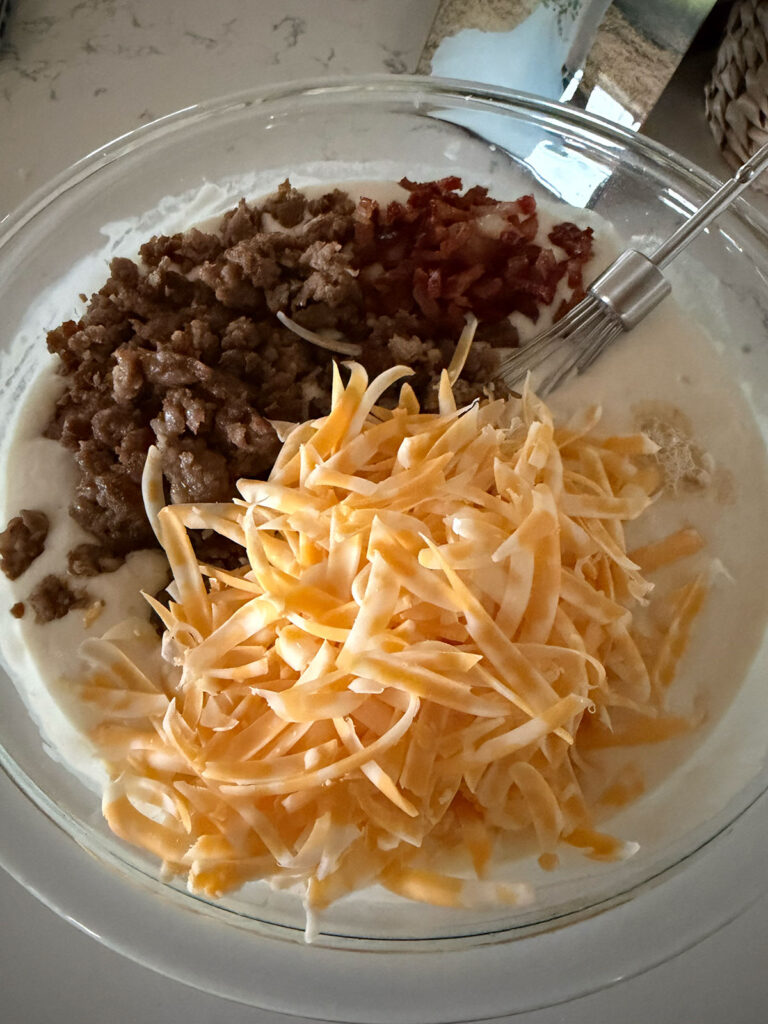 A glass bowl with shredded cheese, cooked sausage, and pancake batter in a glass bowl with a beater resting on the side.