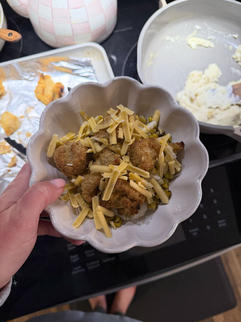 hand holding KFC bowl in white bowl with chicken and mashed potatoes in background