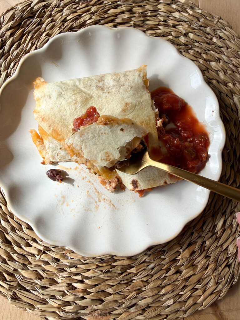 individual serving of sheet pan quesadilla on white plate on placemat. gold fork taking a bite out of quesadilla.