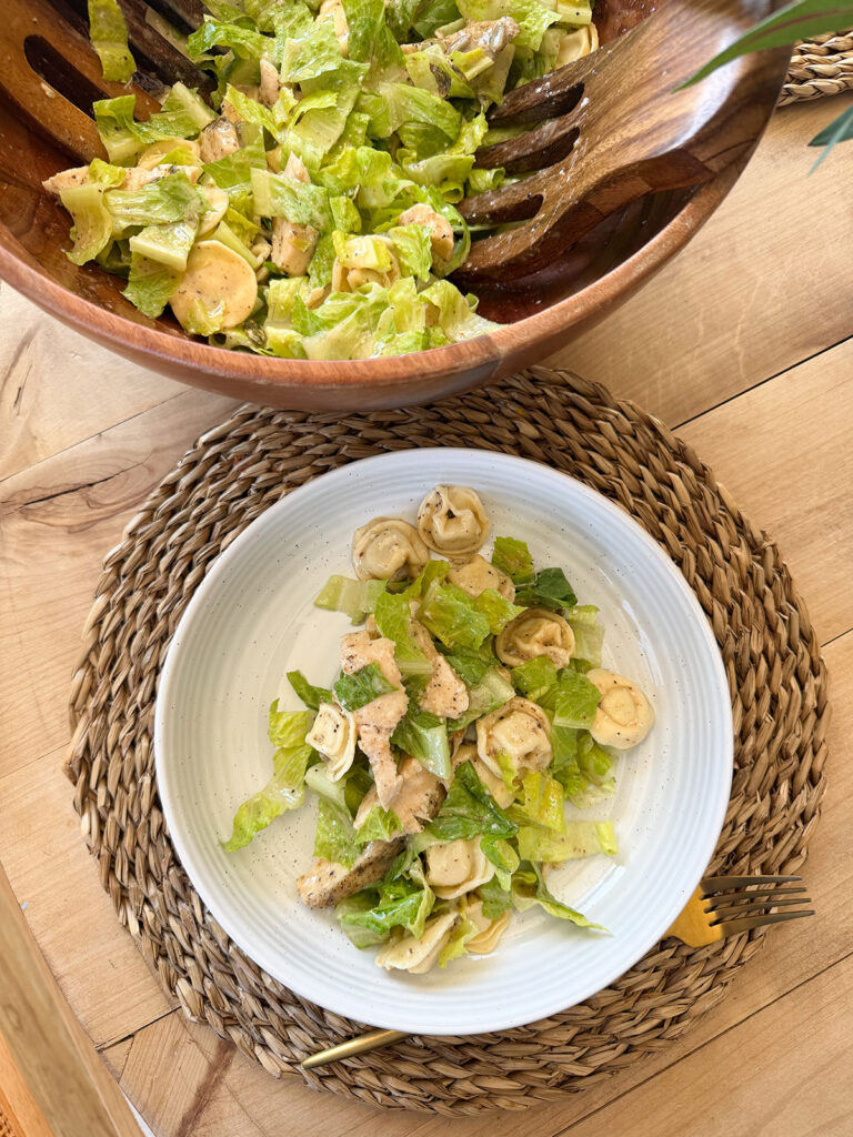 chicken tortellini caesar salad on white plate on placemat with large bowl of salad next to it