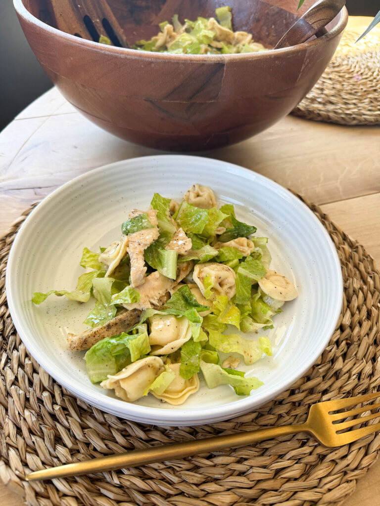 chicken tortellini caesar salad on white plate on placemat with gold fork. Large bowl of salad in background.