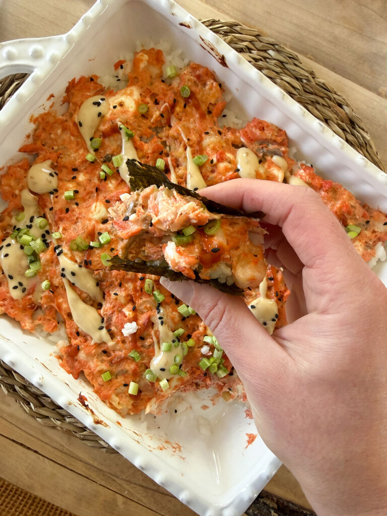 spicy salmon sushi bake in a baking dish with hand holding some sushi bake with nori