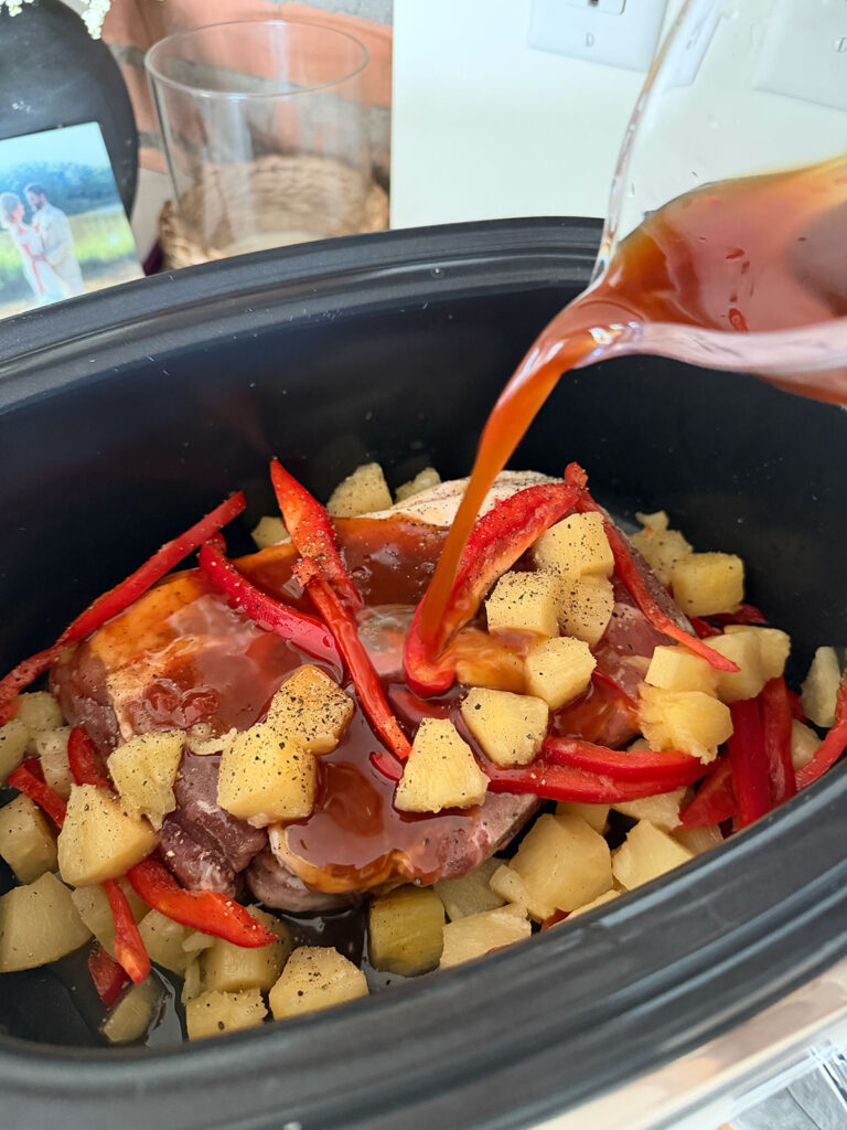 slow cooker sweet and sour pork in base of slow cooker with liquid being poured in over pork, pineapple and bell peppers
