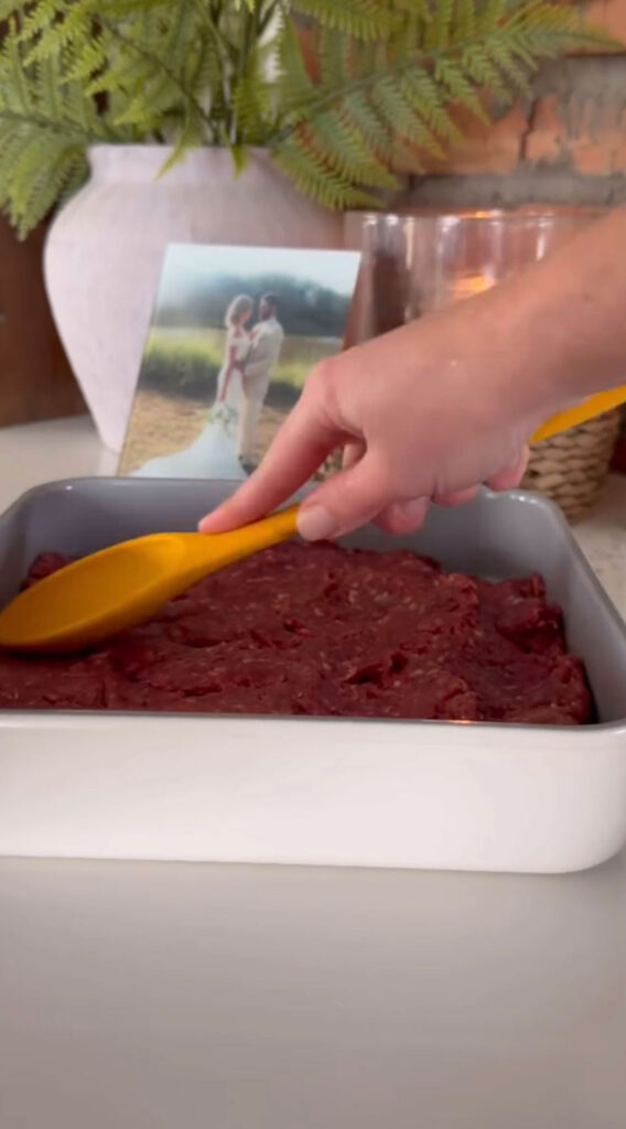 raw meat patty being pressed into pan with hand holding spatula. vase and photo in background.