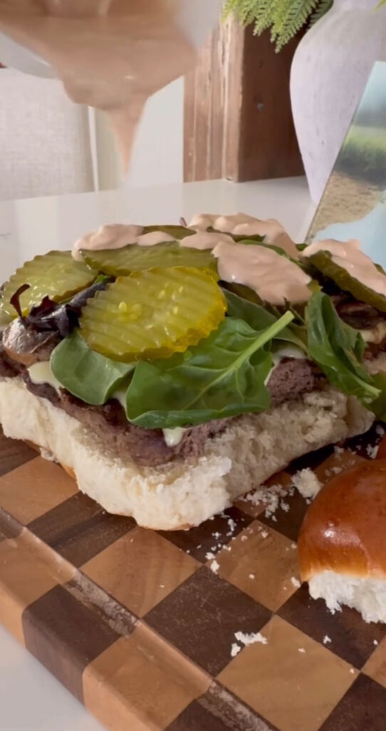 sheet pan burgers being built with meat patty, fresh greens, pickles and burger sauce on top.