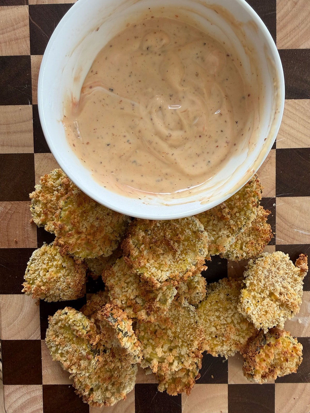 Air fryer dill pickles on a checkered cutting board with a white bowl of dipping sauce.
