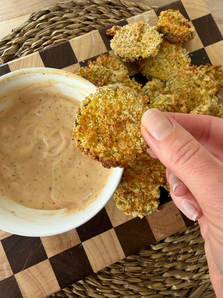 A hand holding an air fryer dill pickle with a bowl of dipping sauce on a checkered cutting board.