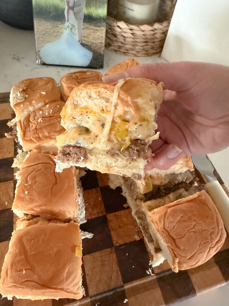 Hand holding one sheet pan burger, side view. Rest of sheet pan burgers on checkered cutting board in background.