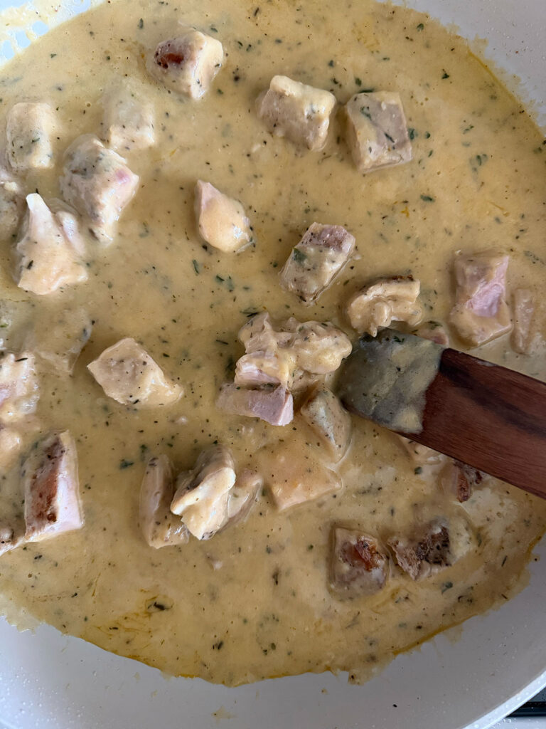 Sauce cooking in a white pan with chicken cubes and a wooden spoon. 