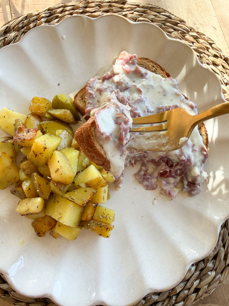 gold fork taking a bite from cream chipped beef on toast, potatoes next to it on white scalloped edge plate