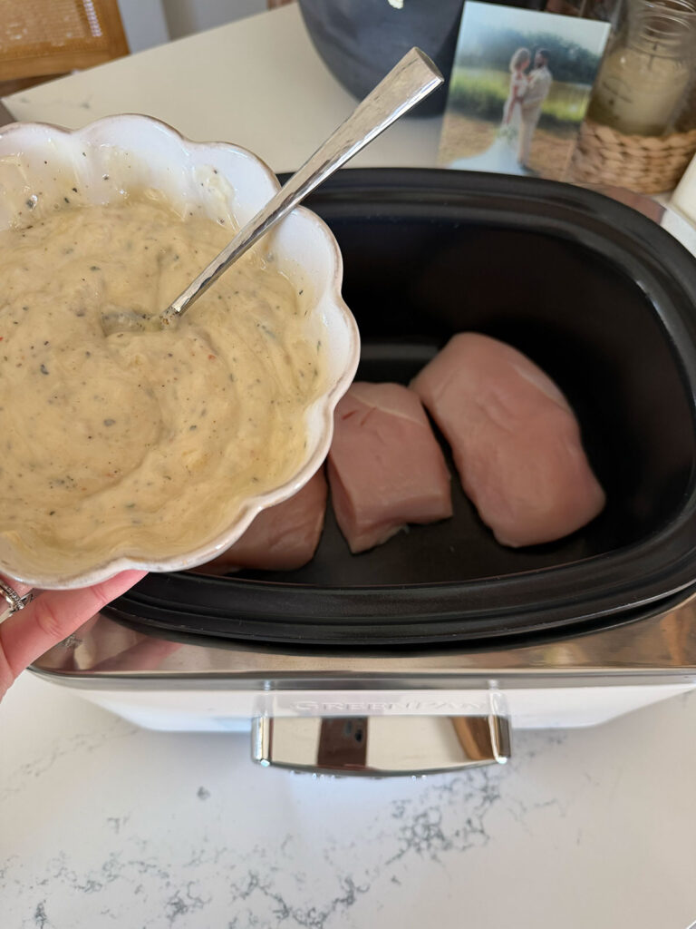 white bowl of cream mixture with spoon in it above raw chicken in slow cooker