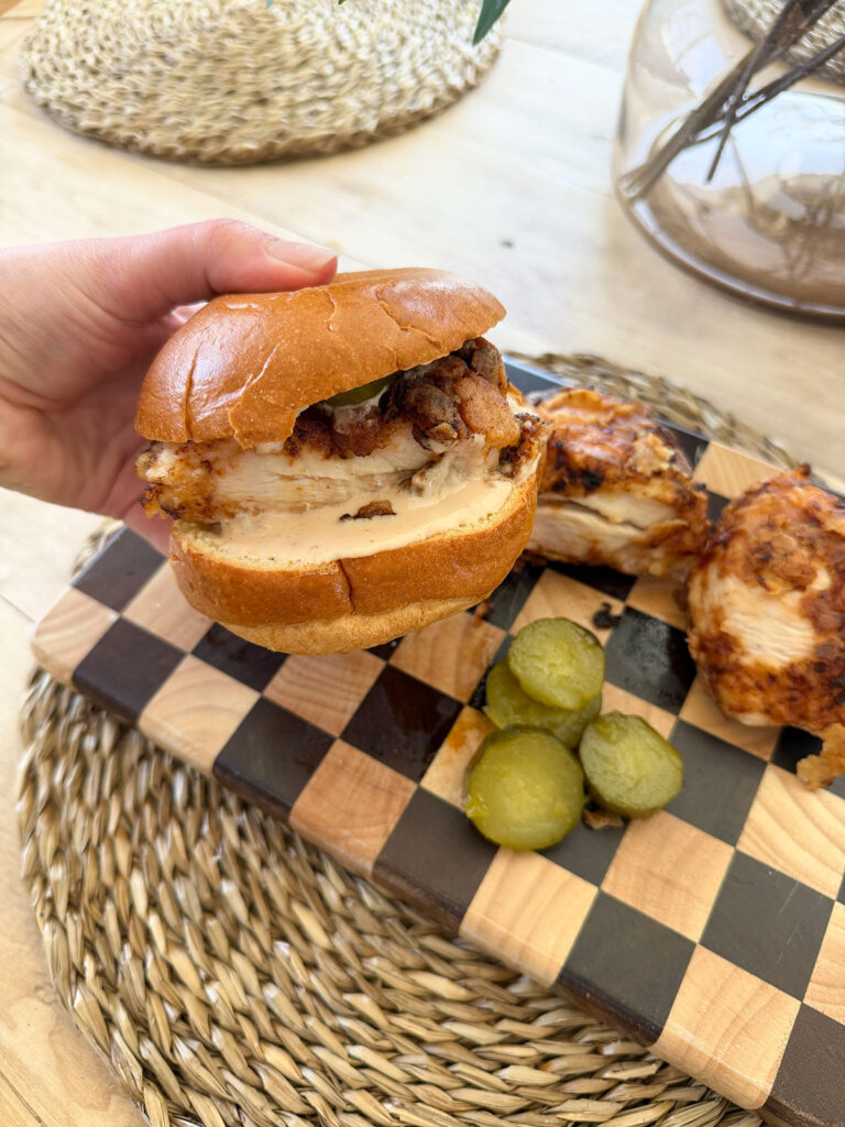 hand holding fried chicken sandwich with friend chicken pieces and sliced pickles in background on checkered cutting board on placemat