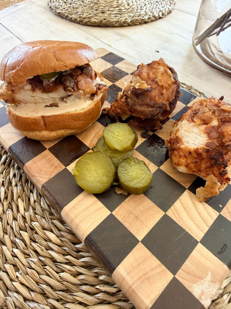 fried chicken sandwich next to two other fried chicken pieces and sliced pickles on checkered cutting board, on placemat on table