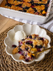 A small white bowl of peach blueberry cobbler with a baking dish of it in the background.