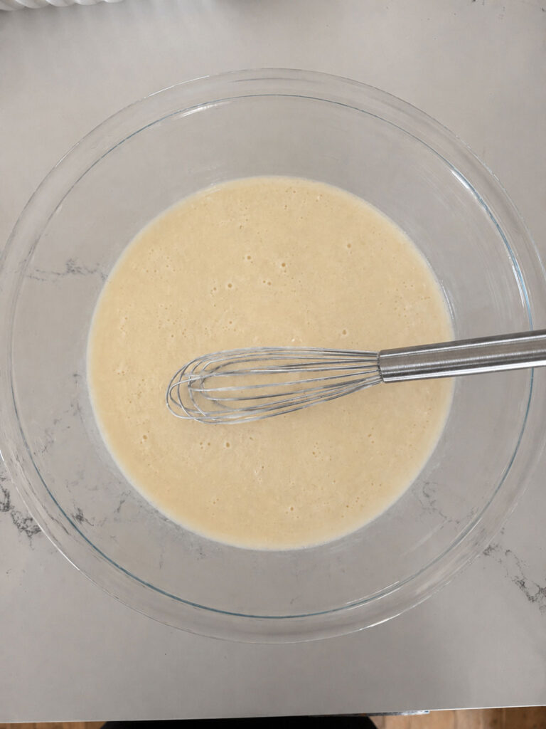 A glass mixing bowl with batter for peach blueberry cobbler with a whisk.