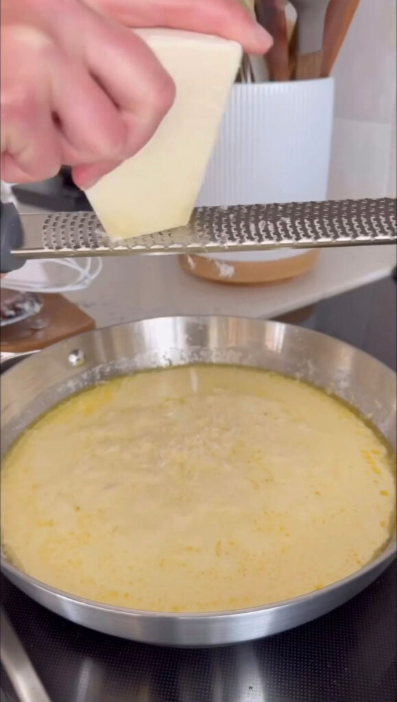 hand grating block of cheese with rasp into alfredo sauce in pan