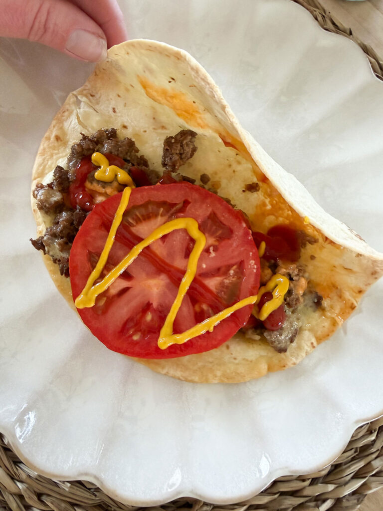 cheeseburger taco on white plate and hand opening it to show slice of tomatoe and drizzle of mustard