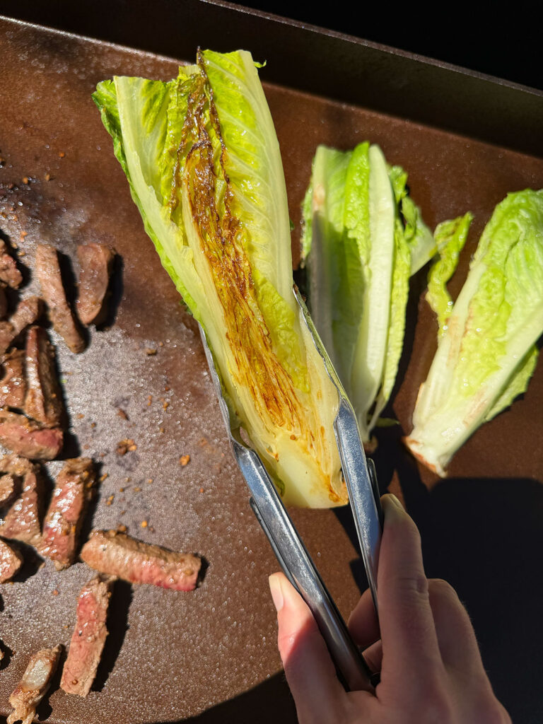 Hand with tongs holding grilled romaine that's charred. More romaine and steak on grill in background.