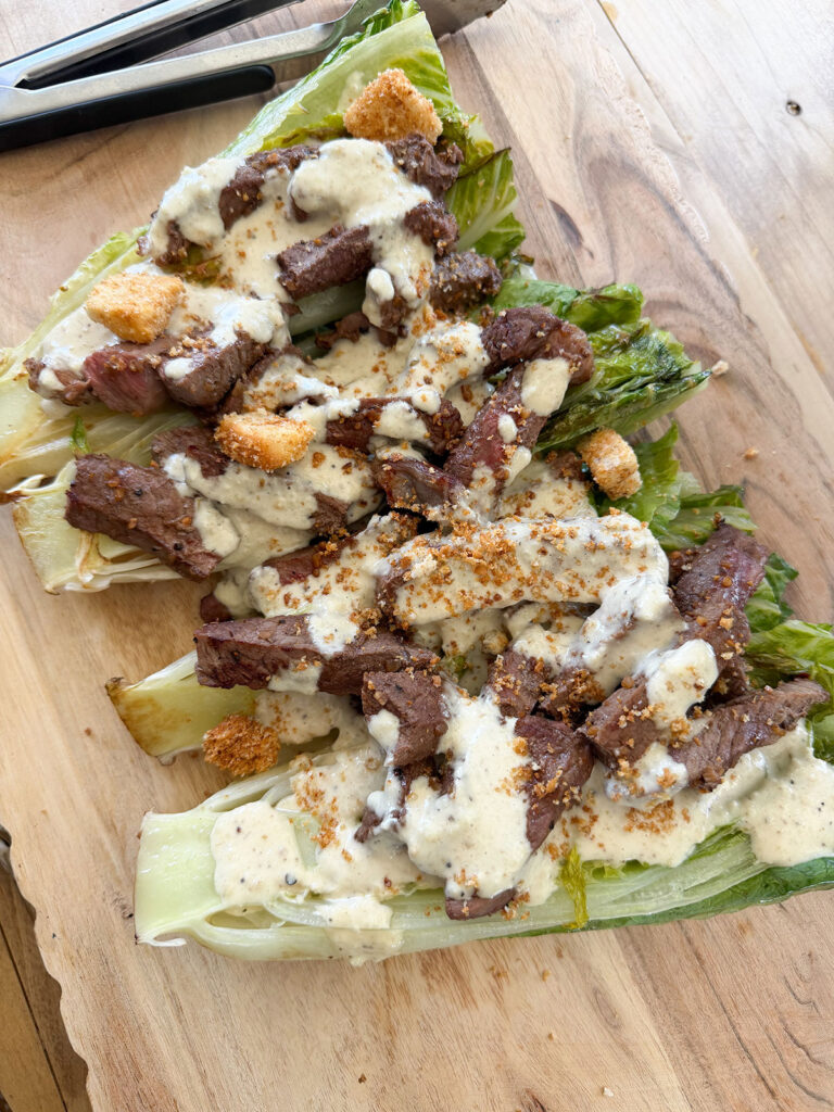 grilled romaine topped with steak, croutons and caesar dressing on wooden cutting board next to tongs