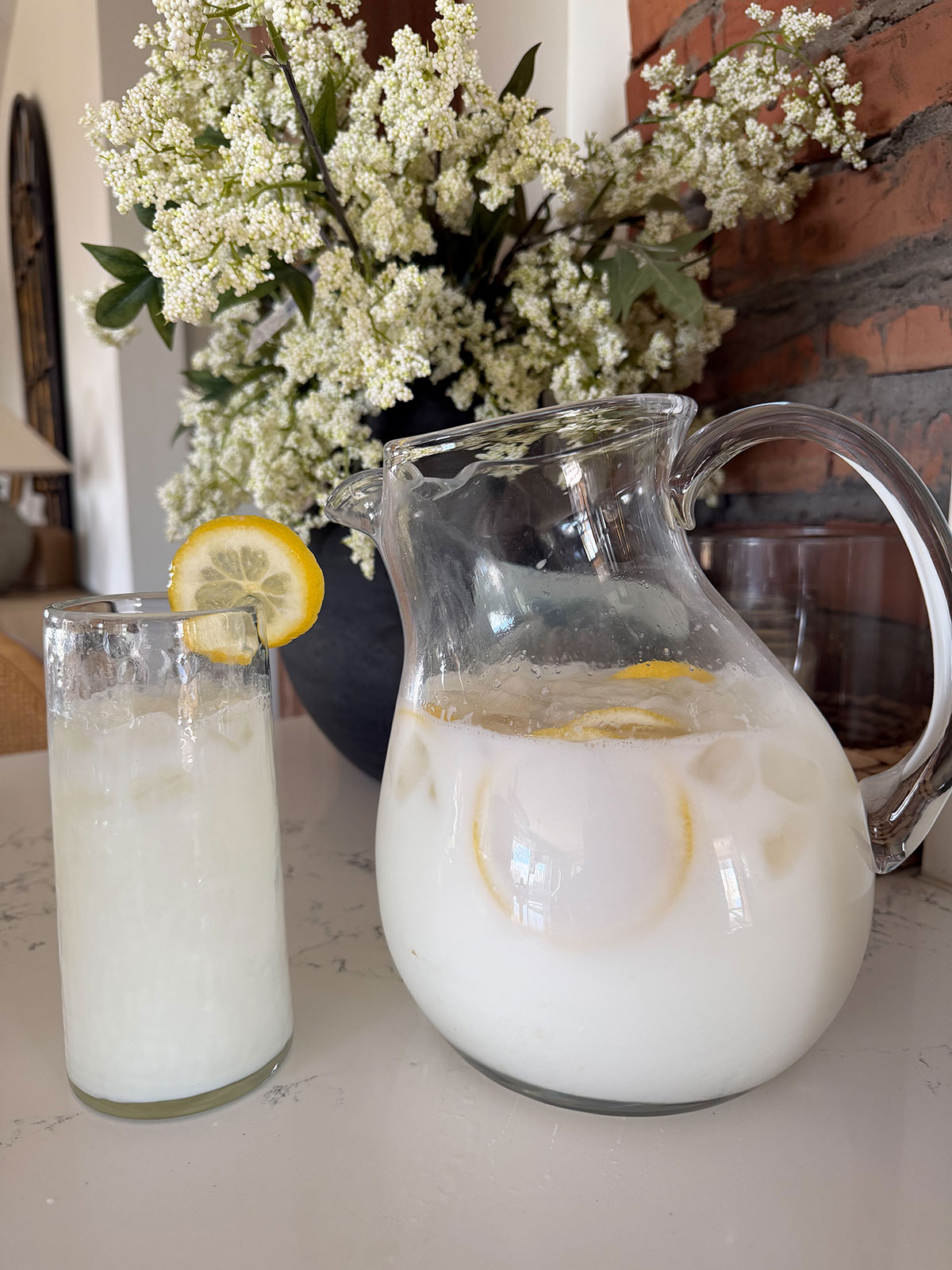pitcher of creamy lemonade next to a glass of creamy lemonade. flowers in background.