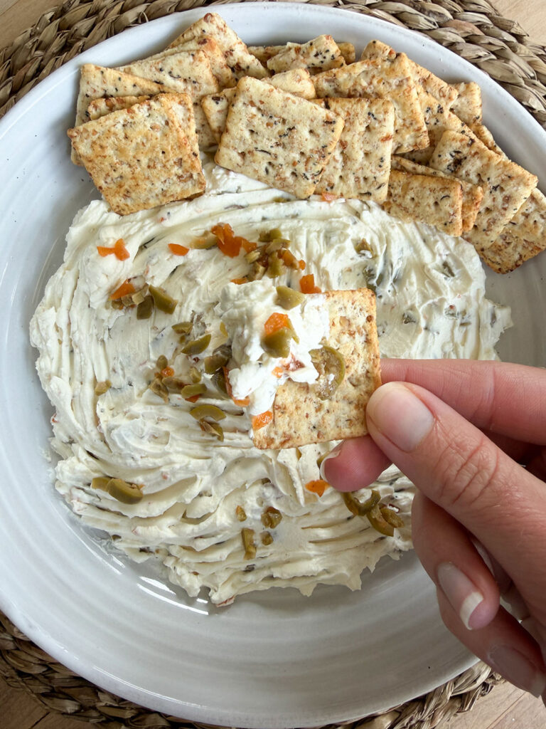 finished olive dip in white bowl with crackers next to it. hand holding cracker dipped into dip.