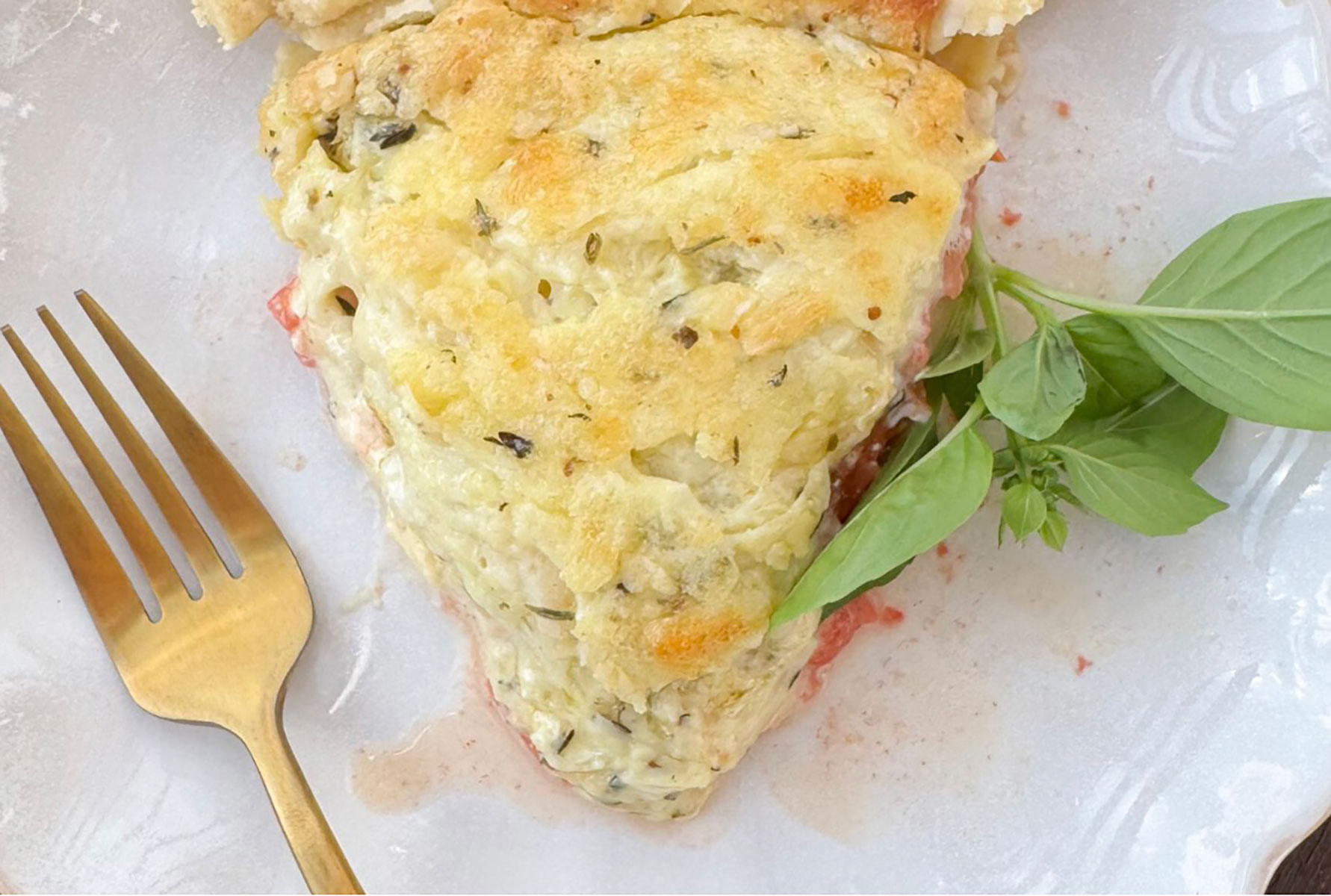 Baked slice of tomato pie on white plate with gold fork and sprig of fresh basil
