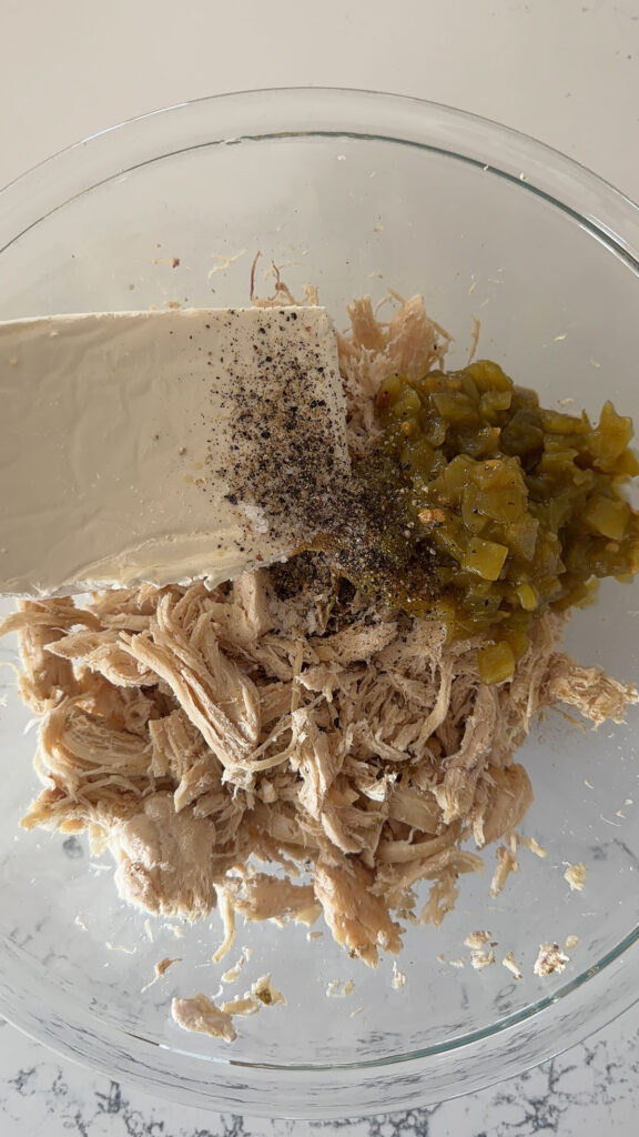 A glass bowl with shredded chicken, green chilies, cream cheese and pepper.
