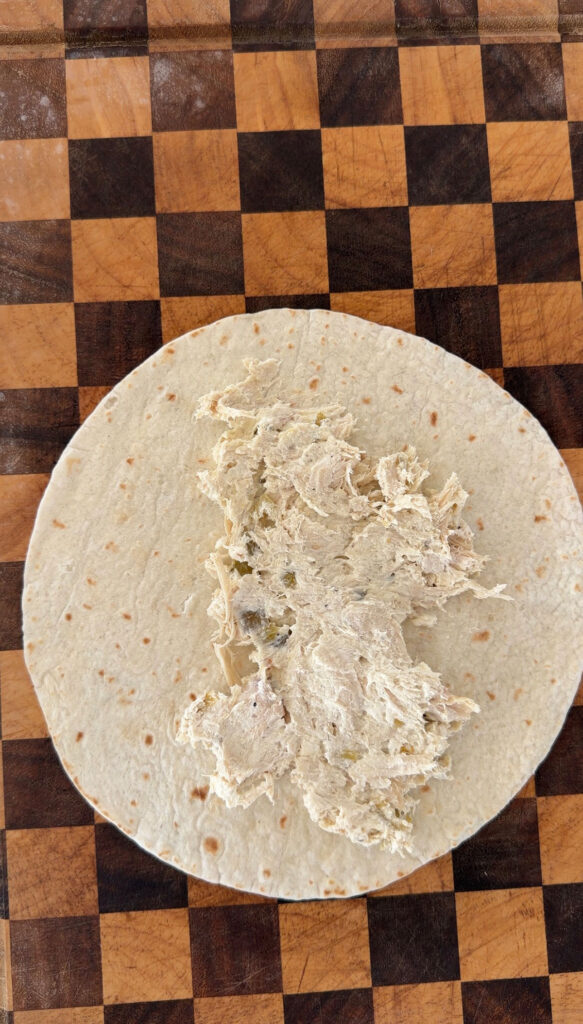 A checkered cutting board with a flour tortilla and chicken mixture inside for enchiladas.