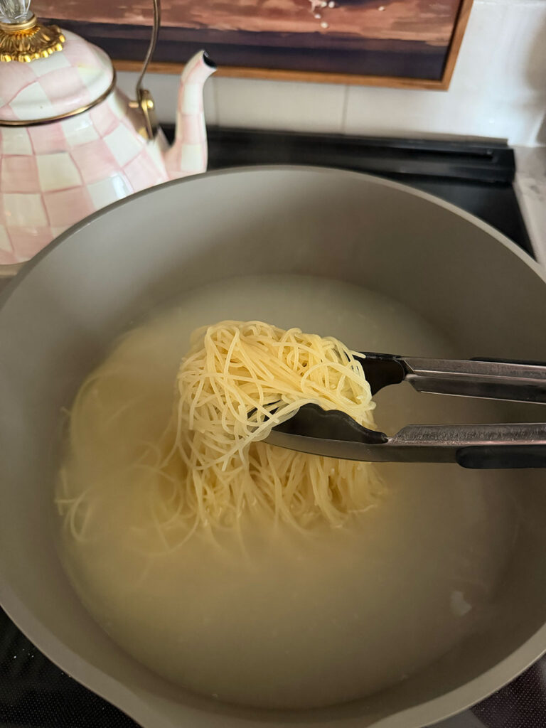 pasta cooking in water, being pulled out with tongs. kettle in background.