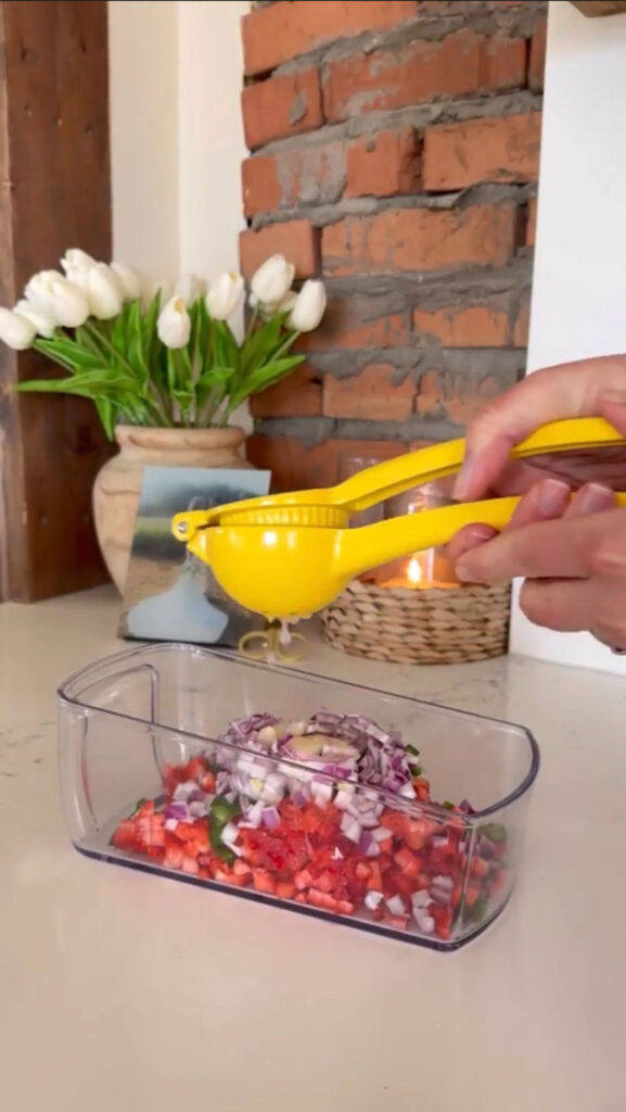 A hand squeezing a citrus press with lime and a chopper with chopped vegetables underneath.